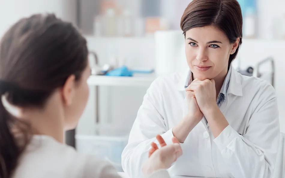 Eine weibliche Person ist mit dem Rücken zur Front und eine lächelnde weibliche Person sitzt vor Ihr mit dem Kopf gestützt auf beide Hand. Die Arme sind auf einen Tisch gestützt