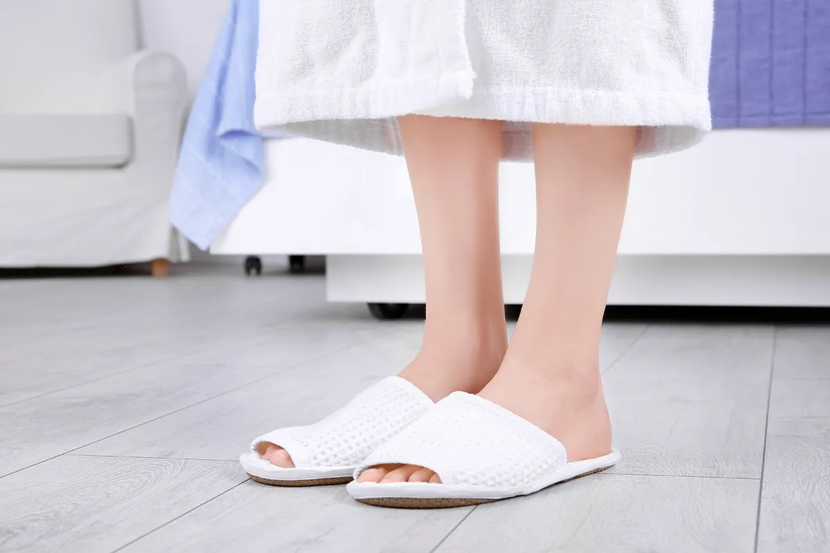 Pilzinfektion Ursachen im Überblick Eine Person in Bademantel und -schlappen steht auf einem gekacheltem Boden und man sieht bis knapp unter die Knie