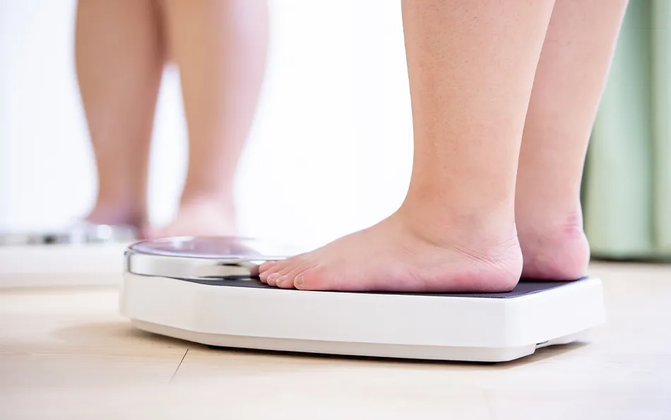 Pilzinfektion Ursachen Risiken Übergewicht Jemand mit dickeren Beinen vor einem Spiegel auf einer Waage