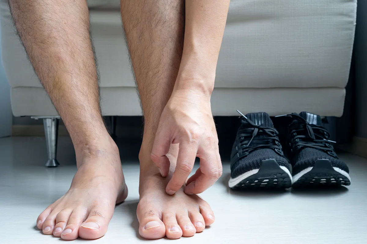 Eine männliche Person hat die Fingernägel auf dem linken Fussrücken angesetzt zum kratzen. Daneben stehen Sportschuhe